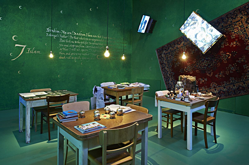 Ein Raum mit Teppich und Bildprojektionen an der Wand, in dem vier Tische stehen, darauf Bücher, drumherum Stühle