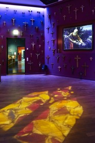 Blick in einen Raum mit vielen Kruzifixen an der Wand und einer Bildprojektion mit Darstellung der biblischen Geschichte