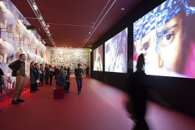 Besucher in einem Ausstellungsraum, rechts an der Wand weiße Skulpturen von Schafen, rechts eine Videoprojektion
