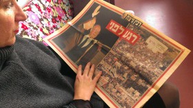 Eine Frau hält eine aufgeschlagene Zeitung in der Hand, auf der ein Bild von Yitzchak Rabin und eines von der Friedenskundgebung abgedruckt ist.