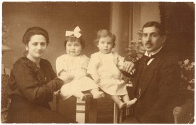 Schwarz-weiß Fotografie: Links sitzt eine Frau in dunklem Kleid, rechts ein Mann im Anzug. Zwischen ihnen sitzen auf einem Tisch zwei Kleidkinder in hellen Kleidern.