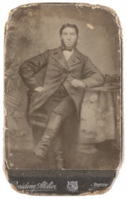 Schwarz-weiß Fotografie eines Mannes in Gehrock und Stiefeln, der an einem Tisch sitzt