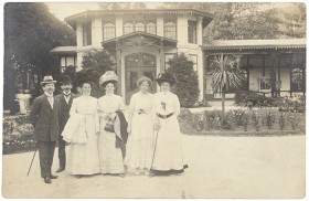 Schwarz-weiß Fotografie: Zwei Männer im Anzug und mit Hut und vier Frauen in hellen Kleidern stehen in einem Garten vor einem Haus