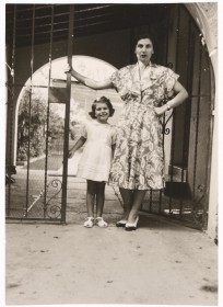 Schwarz-weiß Fotografie: Eine Frau im Kleid und ein siebenjähriges Mädchen stehen an einem Gittertor