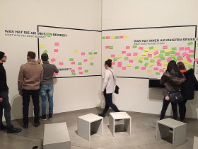 Drei Besucher und drei Besucherinnen stehen vor einer Wand, an die bunte Post-its geklebt sind.