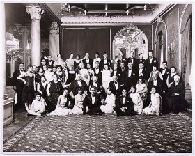 Schwarz-weiß Fotografie con etwa 50 festlich gekleideten Personen in einem prunkvollen Saal