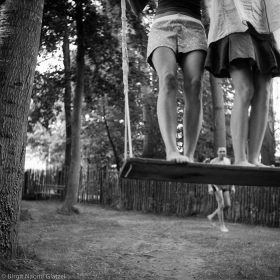 Schwarzweiß Fotografie mit den Beinen von zwei Frauen auf einer Schaukel, im Hintergrund ein Mann