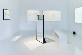 Ein weißgestrichener Raum mit Zeichnungen an den Wänden und einem Spiegel in der Raummitte