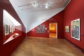 Ausstellungsraum mit roten Wänden, an denen Bilder hängen oder in die Vitrinen eingelassen sind