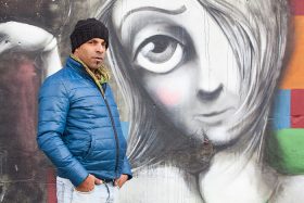 Ein Mann mit Mütze und warmer Jacke vor einer Wand mit dem Grafitti eines Frauengesichts