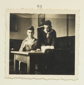 Zwei Jungen an einem Tisch, einer sitzend mit einer Tasse, einer schräg hinter ihm stehend (Schwarz-Weiß-Fo­to)