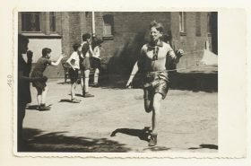 Im Bildvordergrund ist ein rennender Junge zu sehen, der im Begriff ist, die Ziellinie zu erreichen, im Bildhintergrund zuschauende Jungs (Schwarz-Weiß-Fo­to)