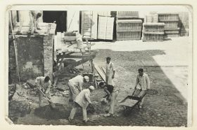 Sieben Männer in Arbeitskleidung und mit Schubkarre, Schaufeln und weiterem Arbeitsgerät (Schwarz-Weiß-Foto)