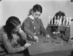 Black-and-white photograph showing two boys and a girl playing dreidel at a table. A Hanukkah menorah stands on the table with all of the candles lit.