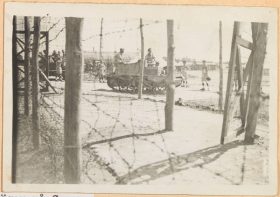 Im Vordergrund des Schwarzweiß-Fotos ist ein Stacheldrahtzaun zu sehen, der an Holzpfeilern befestigt ist. Im Hintergrund zwei Panzer, auf denen Soldaten sitzen, darum herum stehen vereinzelt Internierte.