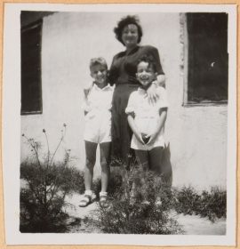 Auf der schwarz-weiß Fotografie legt Leonie im dunklen Kleid ihre Hände auf die Schultern der links und rechts stehenden und in weißen Shorts und Hemden gekleideten Kinder. Im Hintergrund ist eine Hauswand zu sehen.