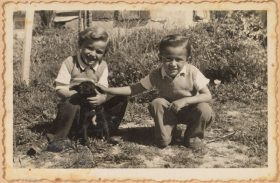 Auf dem schwarz-weiß Bild hocken die beiden Kinder in einem Garten und streicheln einen Hundewelpen.