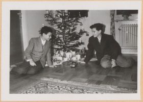 Auf dem schwarz-weiß Foto sitzen die beiden Jungen vor einem Weihnachtsbaum und blicken lachend auf die Gaben, die darunter drapiert sind.