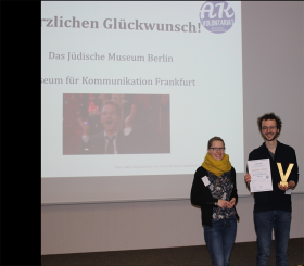 Unter einem Screen, auf dem das AK Volontariat den Gewinnern der Preisverleihung gratuliert, stehen Franziska und David, der die Urkunde und das Goldene V in die Kamera hält.