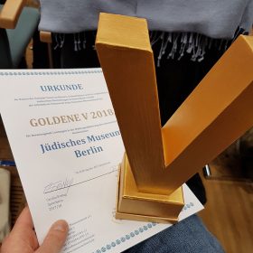 Auf dem Bild ist eine goldene v-förmige Trophäe in Nahaufnahme und die Urkunde zu sehen, auf der das Jüdische Museum Berlin als Gewinner der Auszeichnung vermerkt ist.