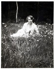 Schwarz-weiß-Fotogafie: Alice liegt in weißem Kleid und mit Kurzhaarschnitt auf einer Blumenwiese.