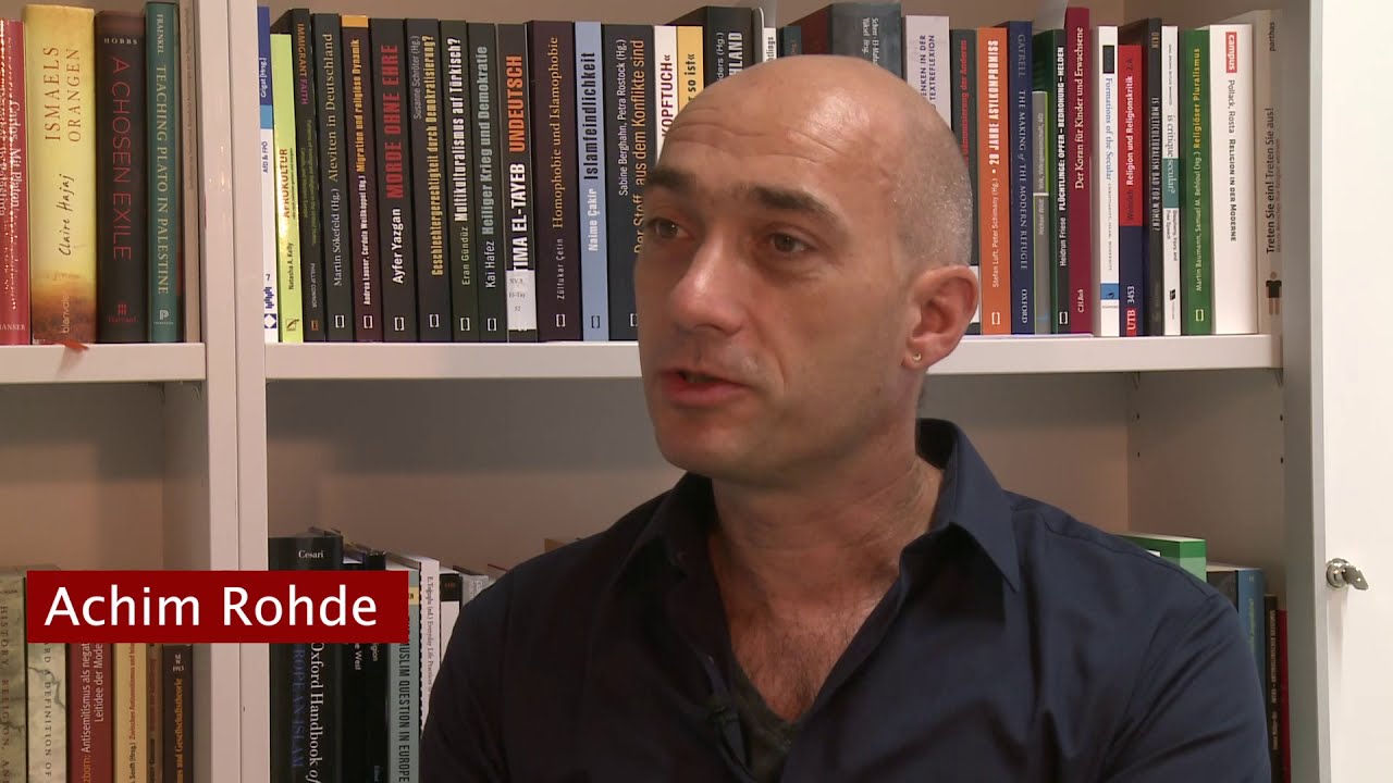 Ein Mann mit schwarzem Hemd sitzt vor einem Bücherregal und gibt ein Interview.