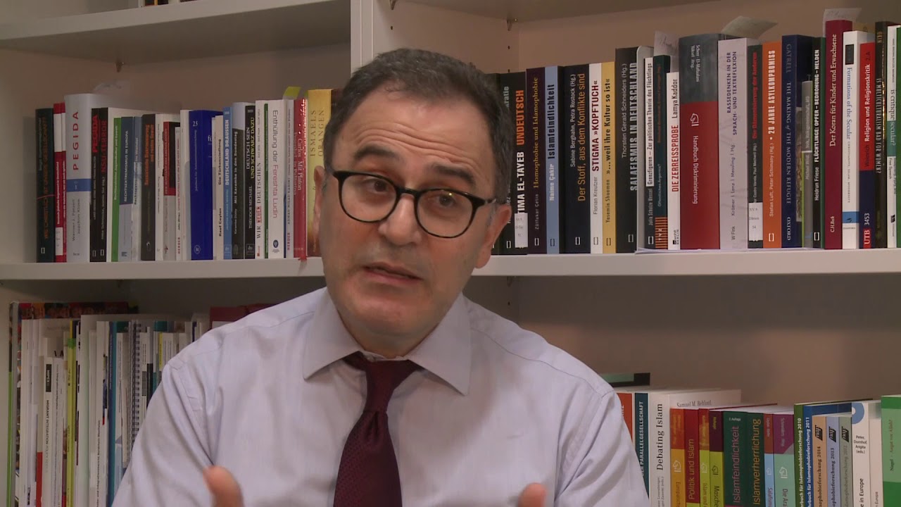 A man with glasses is sitting in front of a bookshelf giving an interview.