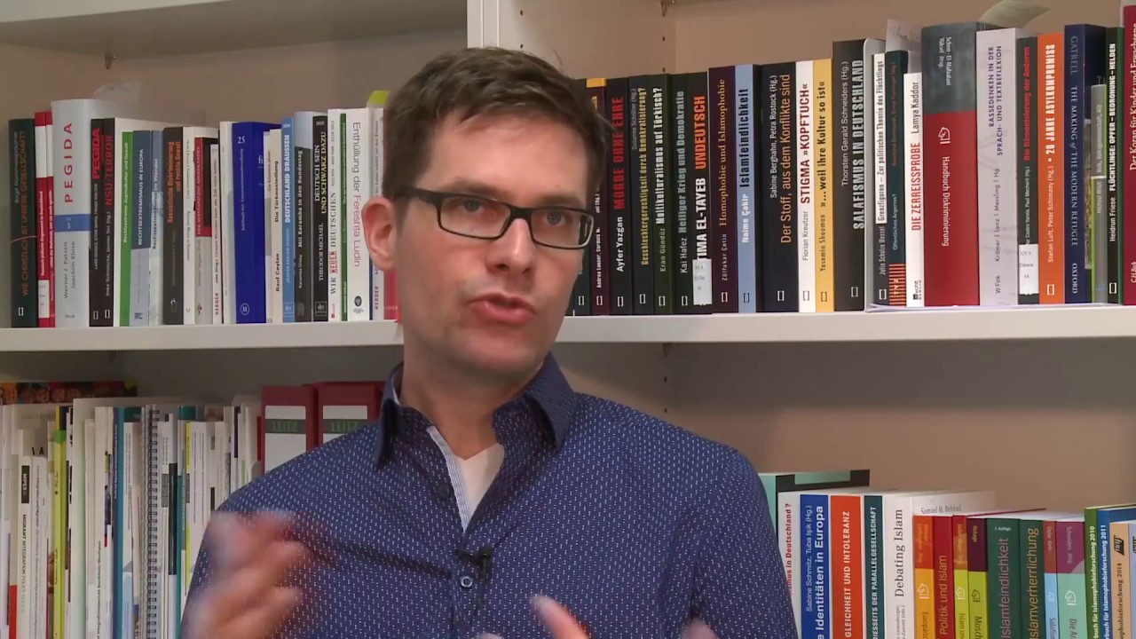 A man with glasses is sitting in front of a bookshelf giving an interview.