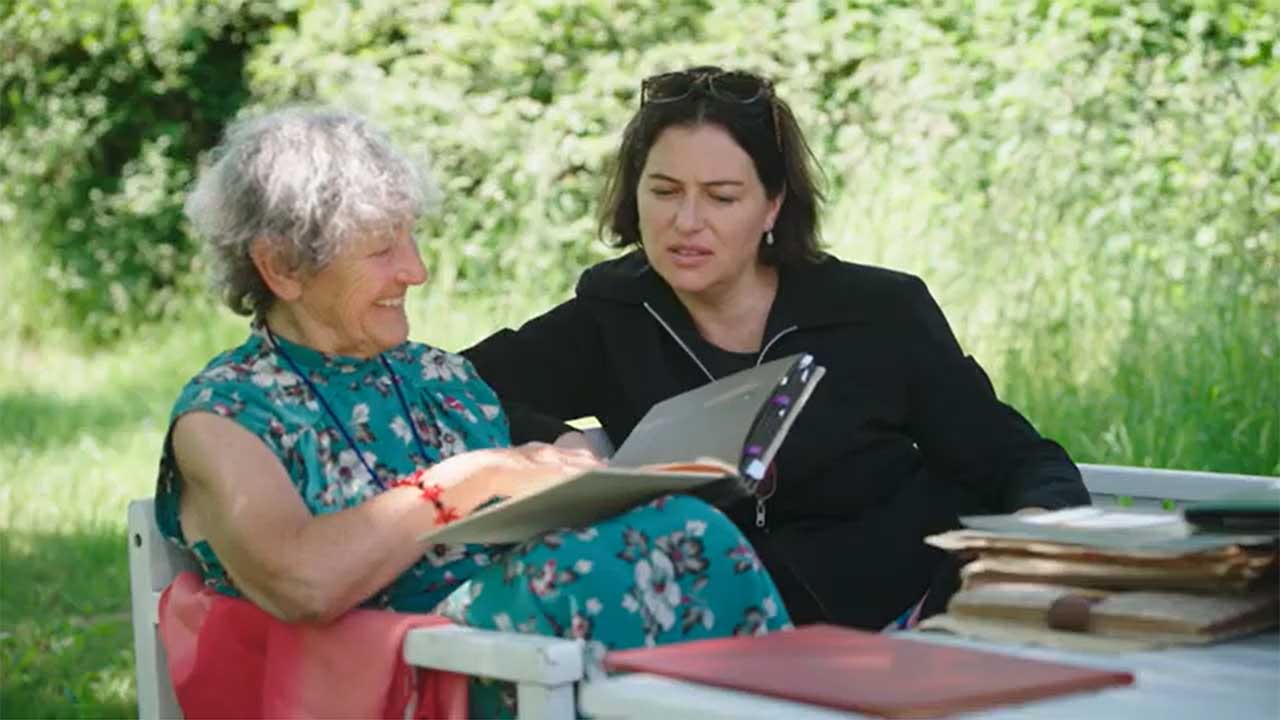 Two women sit on a bench in a garden and look through a photo album together.
