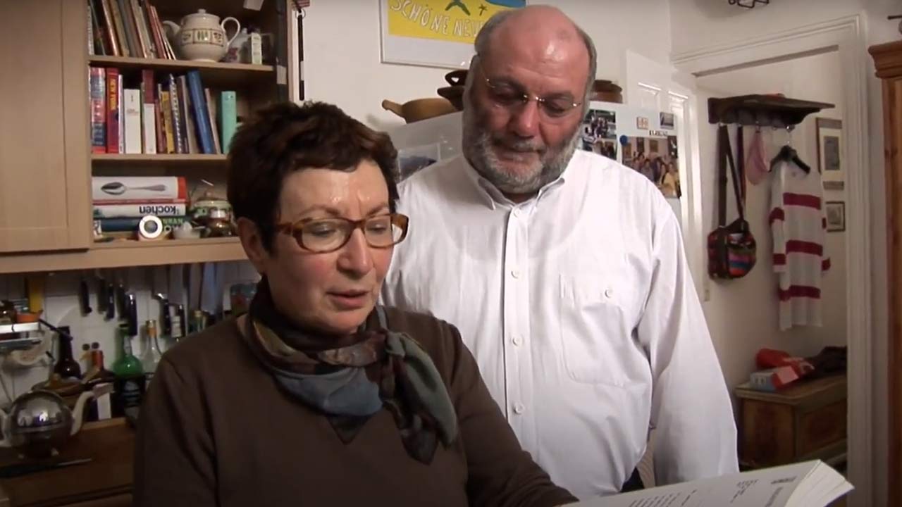 Video still: A woman with short brown hair and glasses shows a book to a bearded man in a white shirt.