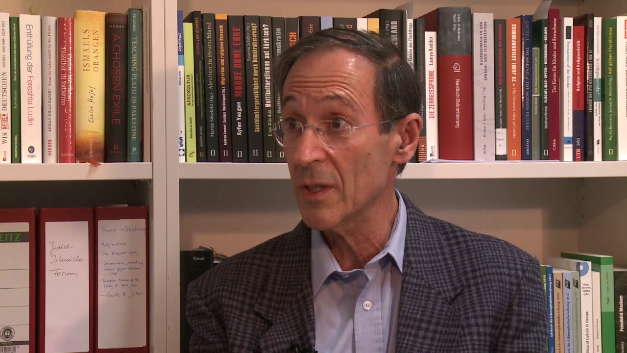 Man with gray jacket sits in front of a bookshelf and gives an interview.
