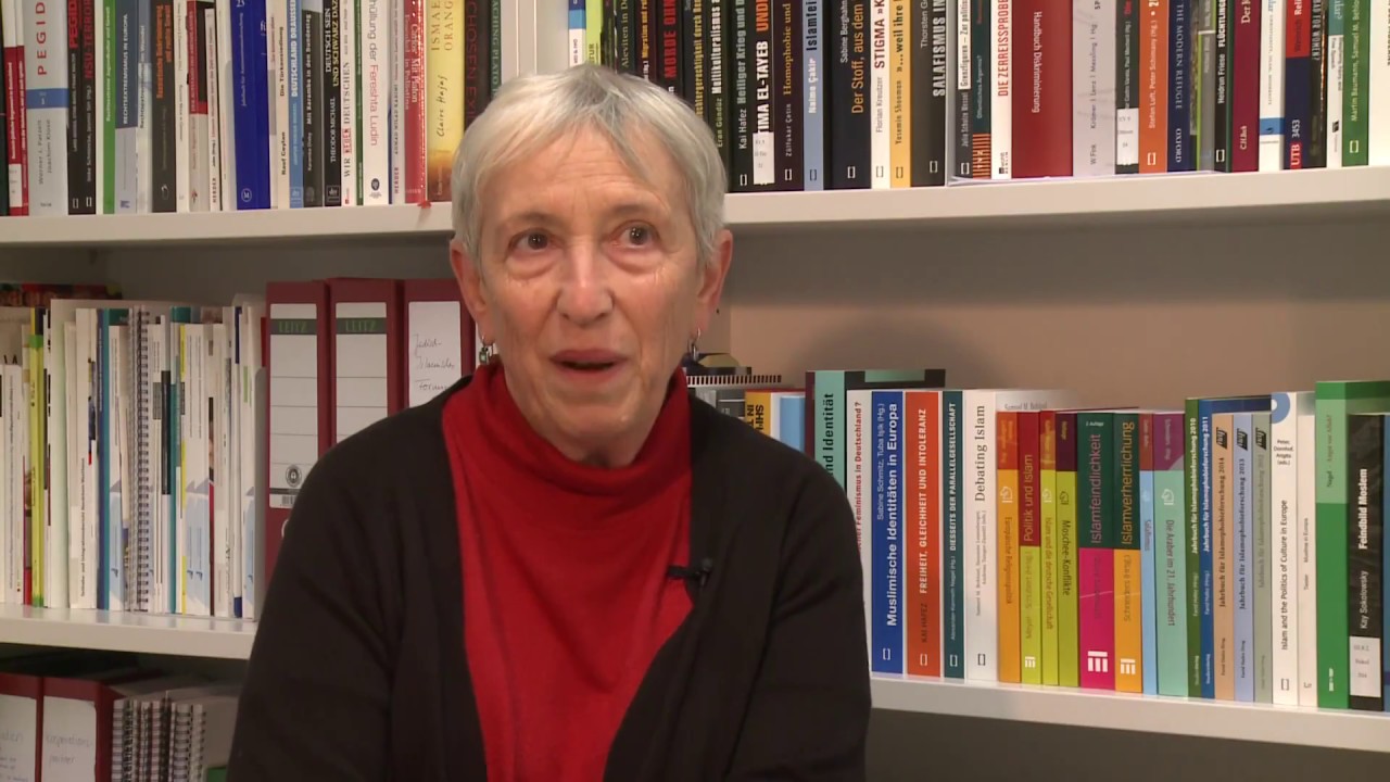 Woman with white hair and black jacket sits in front of a bookshelf and gives an interview.