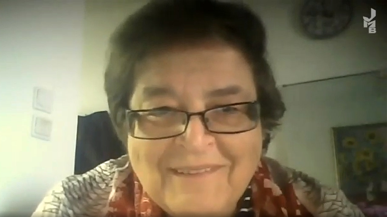 Video still of a smiling elderly woman with glasses.