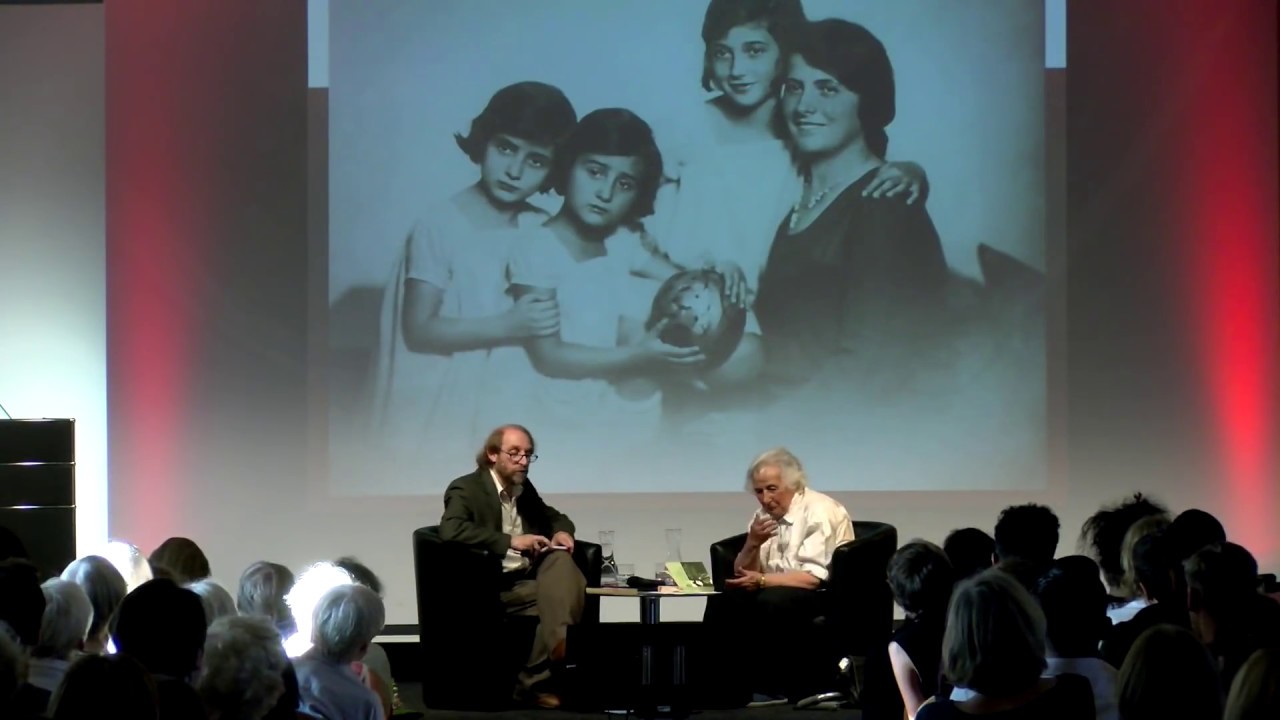 Anita Lasker Wallfisch (right) and Aubrey Pomerance (left) sit on a podium in front of an audience. In the background: projection of a family photo