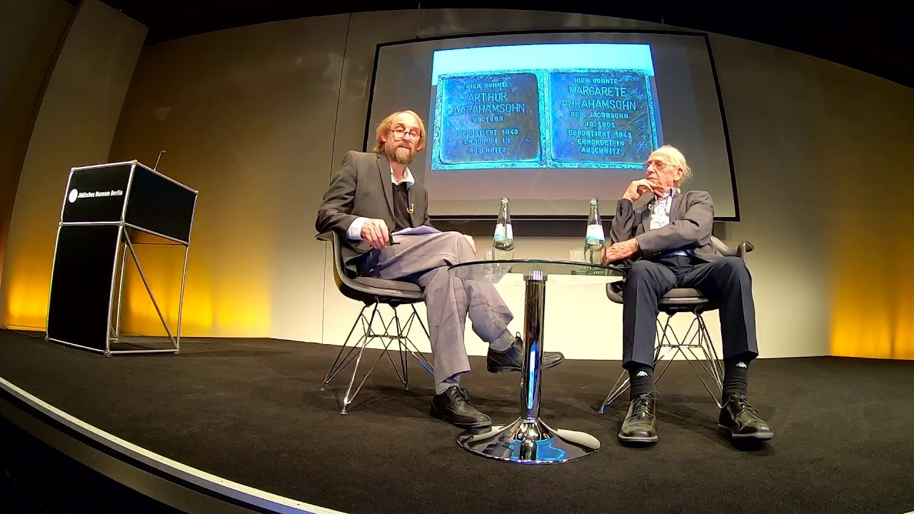 Two men sit talking at a low coffee table: Zvi Aviram on the right, Aubrey Pomerance on the left