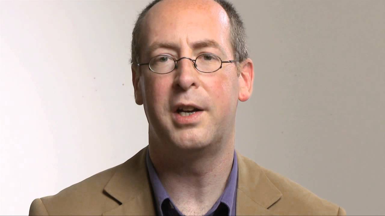 Man with small, round glasses speaks into the camera.
