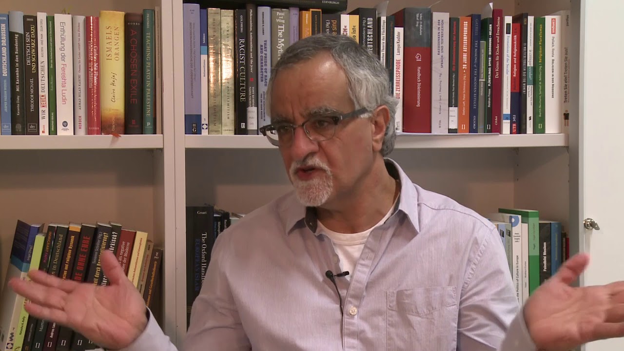A man in a gray shirt sits in front of a bookshelf giving an interview.