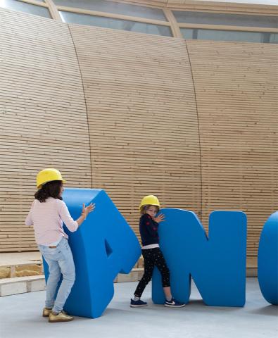 Kinder mit Bauhelmen spielen mit großen Buchstaben des Schriftzugs ANOHA