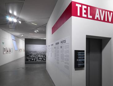 Blick in den Eingangsbereich der Ausstellung: Rechts eine Wand mit Beschriftung, im Hintergrund ein großen Schwarz-Weiß-Foto, das ein Lufbild einer Großstadt zeigt