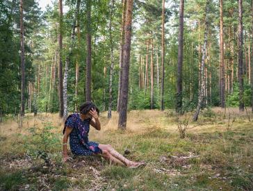Fotografie einer Frau im blauen Sommerkleid, die im Wald auf dem Boden sitzt und sich über der Stirn in die Haare greift