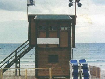 Ein kleines Holzgebäude direkt am Strand, das mit der israelischen Flagge geschmückt ist und von Stapeln von Strandliegen umgeben ist.
