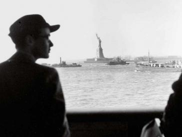 Black and white historical photograph of a man and woman in the foreground, the Statue of Liberty surrounded by boats can be seen in the distantance.