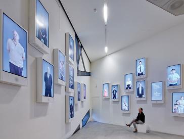 Exhibition space with numerous screens on the walls, each of which shows a person looking at the visitor.