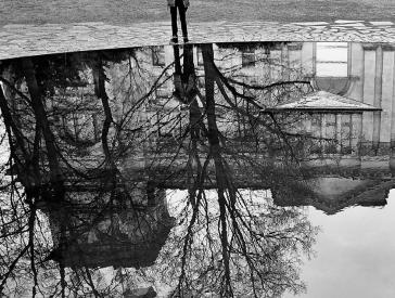 Black and white photograph: a boy stands in front of a puddle, an old building is reflected in the puddle.