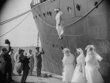 A black and white film still shows a crowd of people in front of a ship. On the left are men in suits, on the right are three women in white dresses. They all look up at a woman in a wedding dress hanging from a rope in front of the ship. 