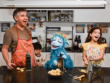 Ein Mann, ein Junge, eine blaue Puppe und ein Mädchen stehen an einer Kücheninsel, reiben Kartoffeln und lachen