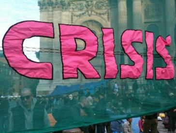 A fabric banner with the words Crisis on it, with people standing in a square behind it.