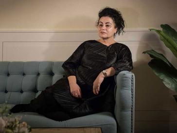 Woman in black dress sits on a sofa.