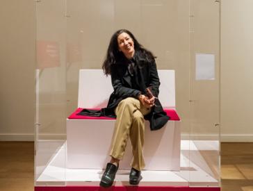 Sharon Adler sitting on a bench in a plexiglass box which is open at the front