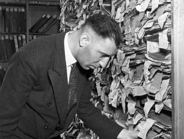 Black-and-white photo of a man in front of a shelving unit overflowing with file folders.
